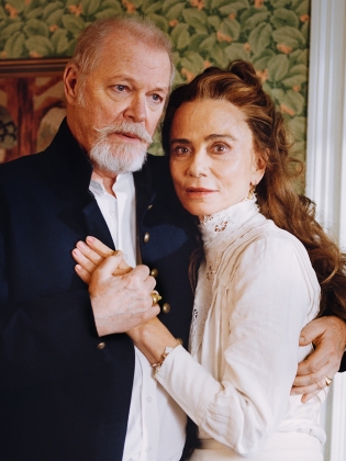 En äldre man med skägg och mörk kavaj håller handen på en kvinna i vit klänning, i ett rum med blommig tapet och tända ljus.