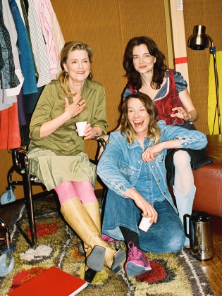 Tre kvinnor sitter i en loge med kläder på galgar. De skrattar och håller i koppar. En termos står på golvet.