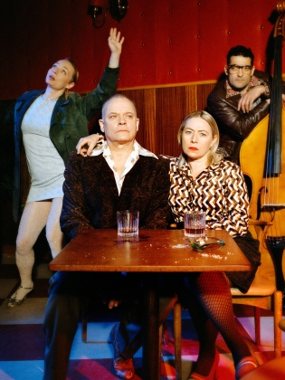 Fyra personer i en bar: ett par sitter vid ett bord med drinkar, en kvinna poserar dramatiskt bakom dem och en man lutar sig mot en kontrabas.