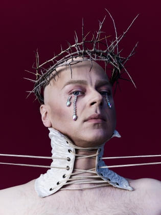 En person bär taggkrona och hög vit snörkrage, kristallprydda tårar rinner nedför kinderna.