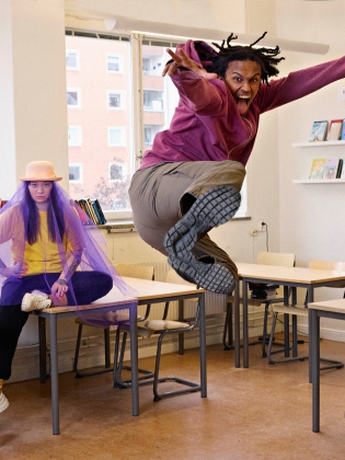 En kille som hoppar i ett klassrum, bakom honom sitter en tjej på ett bord
