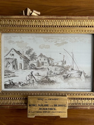 Inramad teckning i svartvitt föreställande ett lantligt hamnområde med hus, människor, båtar och en man till häst i förgrunden. Bilden är omgiven av en rikt dekorerad guldram med en plakett nedtill.