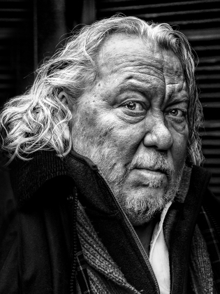 Man med långt, lockigt grått hår och skägg, klädd i halsduk och jacka, ser in i kameran.