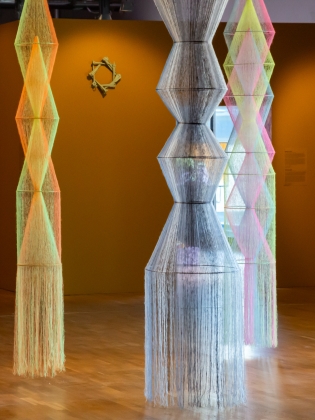 Flera höga, geometriska skulpturer av färgade trådar hänger i ett rum med senapsgula väggar och trägolv.