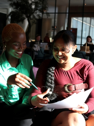 Två unga kvinnor sitter tillsammans, den ena pekar med en penna på papper som den andra håller, båda ler och verkar samarbeta.