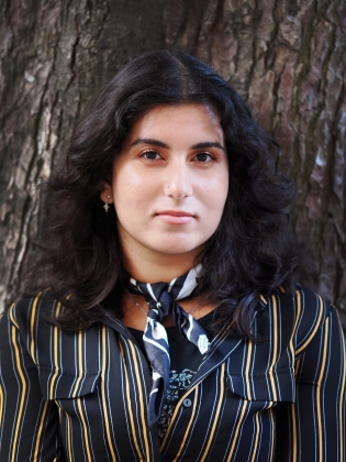Ung kvinna med långt mörkt hår, randig skjorta och scarf står framför ett träd och ser in i kameran.