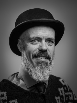 Skäggig man med hatt och islandströja, fotograferad i svartvitt porträtt.