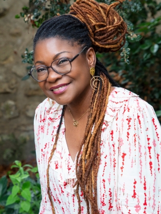 Kvinna med glasögon och långa dreadlocks i vitmönstrad blus, sitter utomhus och ler mot kameran.