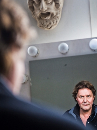 Johan Rabaeus som tittar i en sminkspegel, ovanför spegeln hänger en grekisk teatermask