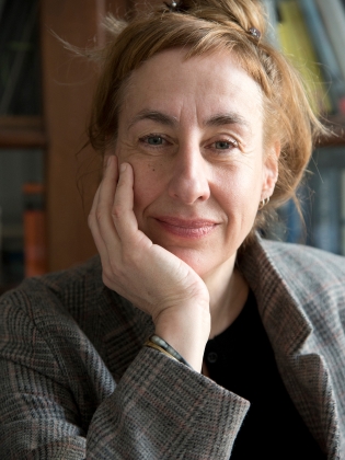 Medelålders kvinna med uppsatt hår och rutig kavaj, lutar huvudet mot handen och ser in i kameran.