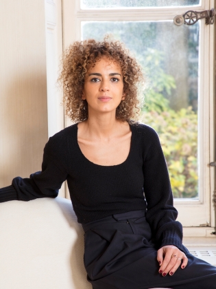 Yngre kvinna med lockigt hår och svart topp sitter inomhus framför ett fönster.