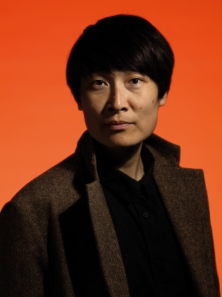 Person med kort, mörkt hår i brun kavaj och svart skjorta, poserar mot orange bakgrund.