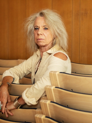 Äldre kvinna med ljust rufsigt hår i beige skjorta sitter i en tom föreläsningssal och tittar mot kameran.