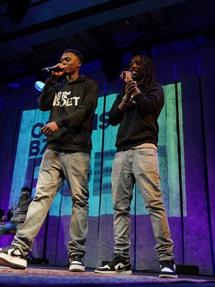 Två unga män i svarta hoodies och jeans uppträder med mikrofoner på scen med färgad bakgrund.