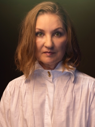 Medelålders kvinna med ljust halvlångt hår i vit skjorta, porträtt mot mörk bakgrund.