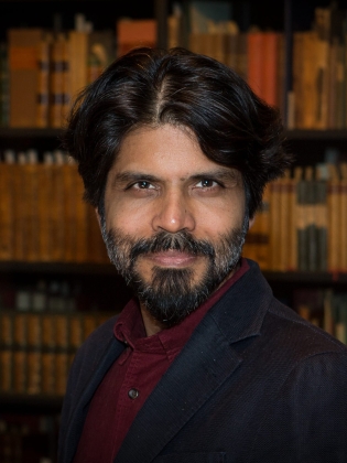 Man med mörkt hår och skägg i vinröd skjorta och kavaj, står framför bokhylla.