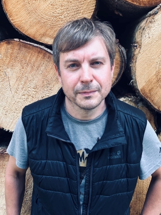 Man med kort ljust hår i grå t-shirt och svart väst, står framför staplade timmerstockar.