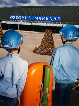 Två pojkar i blå hjälmar och jackor står vända mot varuhuset Domus Marknad, omgivna av blomstertorn.