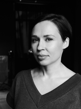 Kvinna med mörkt uppsatt hår i mörk topp, står i svartvitt foto i ett inomhusrum.