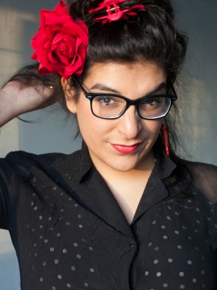 Kvinna med mörkt hår, glasögon och röd hårklämma med stor röd blomma poserar i svart prickig blus.