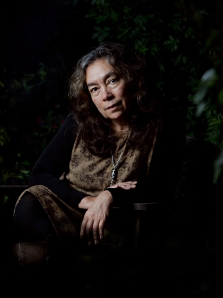 Medelålders kvinna med långt, lockigt hår sitter utomhus i mönstrad klänning, med ena armen vilande på armstödet.