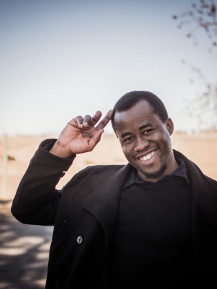 Ung man i mörk kappa och tröja, ler och gör honnör utomhus i lantlig miljö.