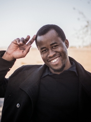 Ung man i mörk kappa och tröja, ler och gör honnör utomhus i lantlig miljö.