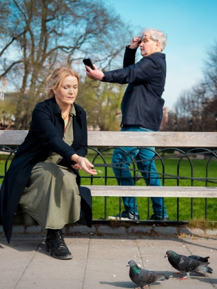 En kvinna som sitter på en bänk och matar duvor, bakom henne står en äldre man och tittar på sin mobil