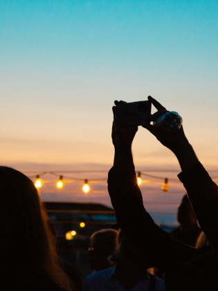 Silhuetter av personer i motljus vid solnedgång, en person håller upp en mobil mot himlen.