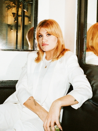 Kvinna med blont hår i vit kostym sitter i hörnbås med spegelreflex bakom sig.